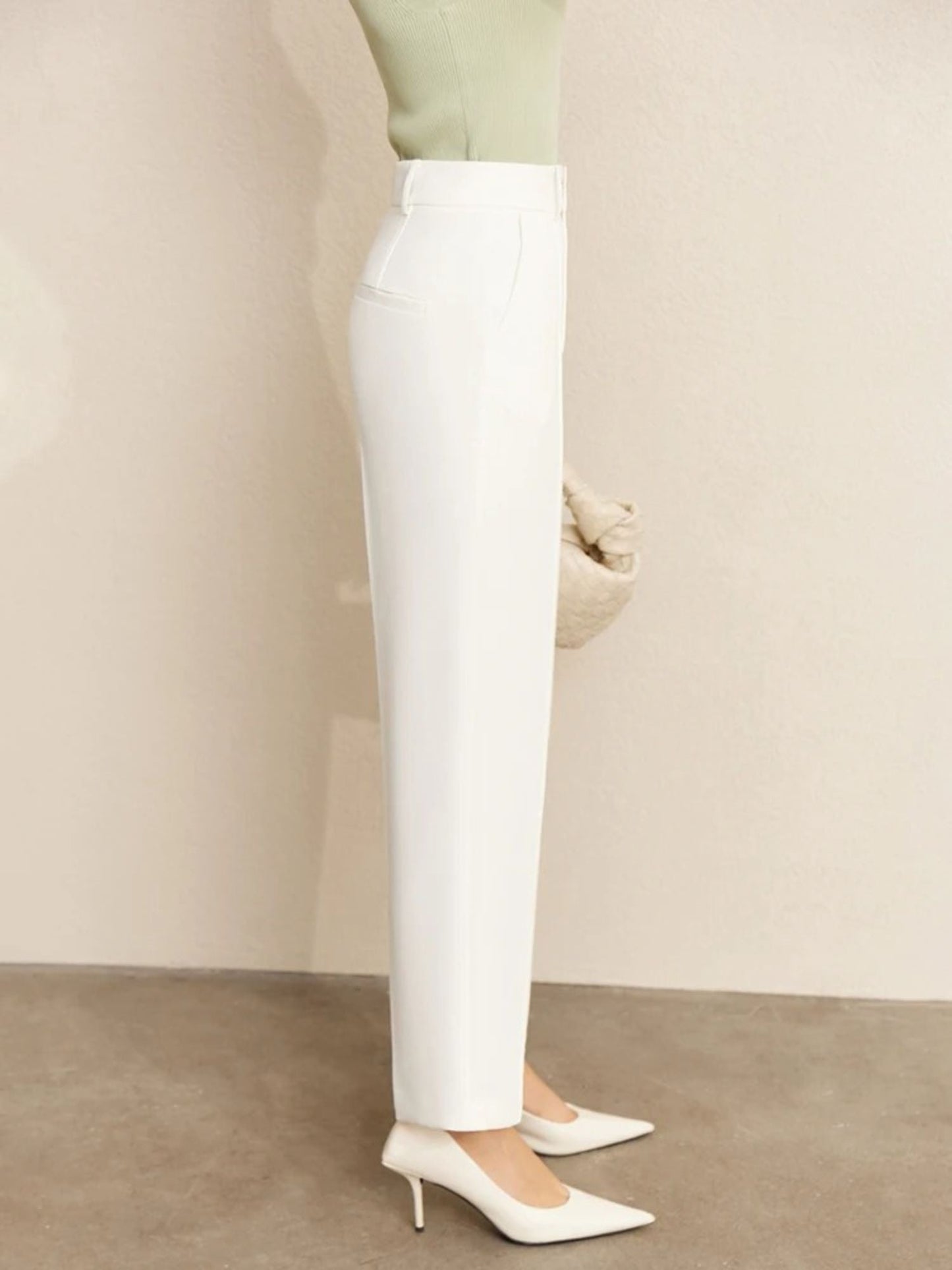Close-up side view of high-waisted white trousers with tucked-in green top and beige handbag