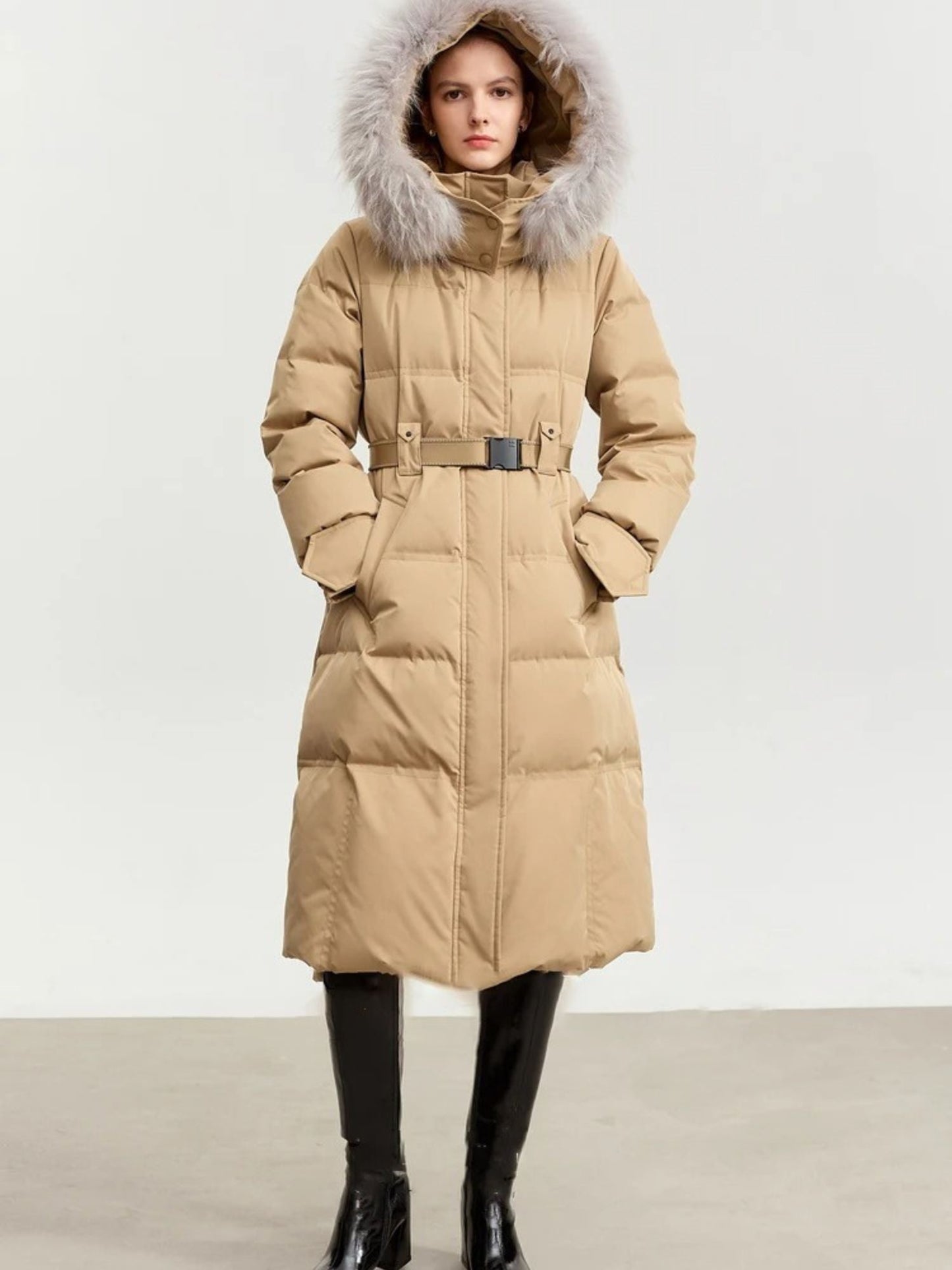 Three-quarter front view of the beige coat, with one hand in the pocket. Belt detail and horizontal quilting are visible
