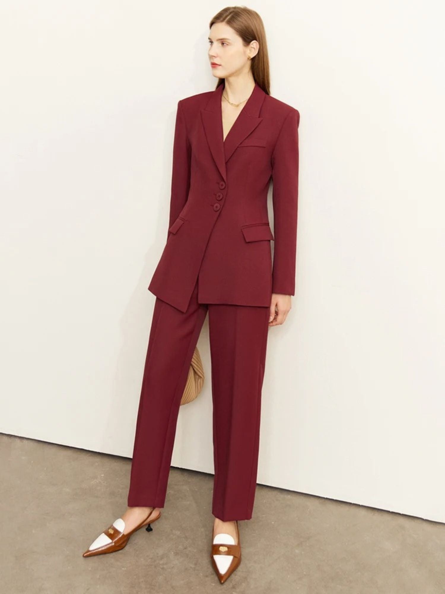 Woman in a deep burgundy tailored blazer and matching trousers, paired with strappy white heels and a beige handbag