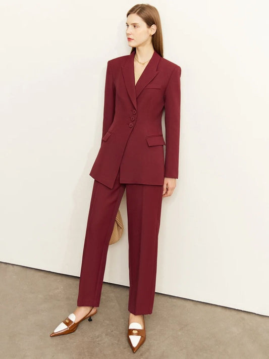 Woman in a deep burgundy tailored blazer and matching trousers, paired with strappy white heels and a beige handbag