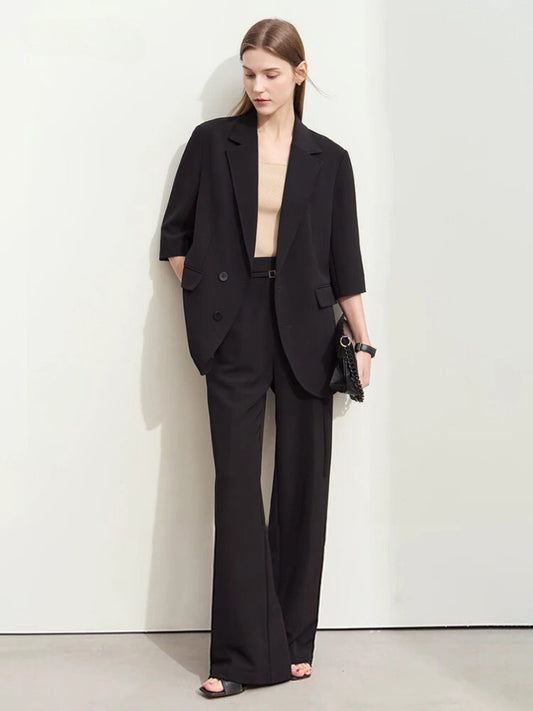 Woman in a black oversized blazer with elbow-length sleeves and matching wide-leg trousers, paired with a beige top and black clutch