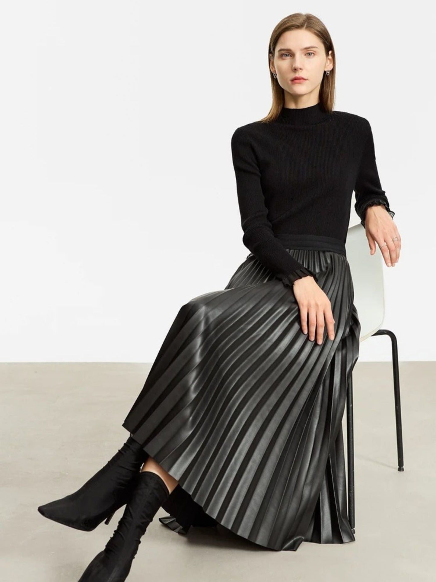 Side view of black faux leather pleated midi skirt worn with black long-sleeve top, model seated