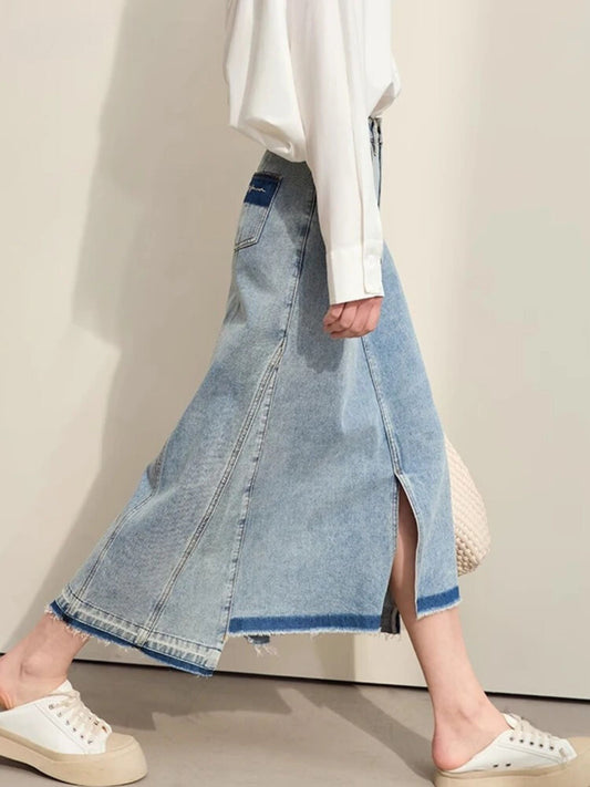 Model wearing a light blue A-line denim midi skirt with front slit and contrast dark hem. Styled with white sweatshirt and white sneakers