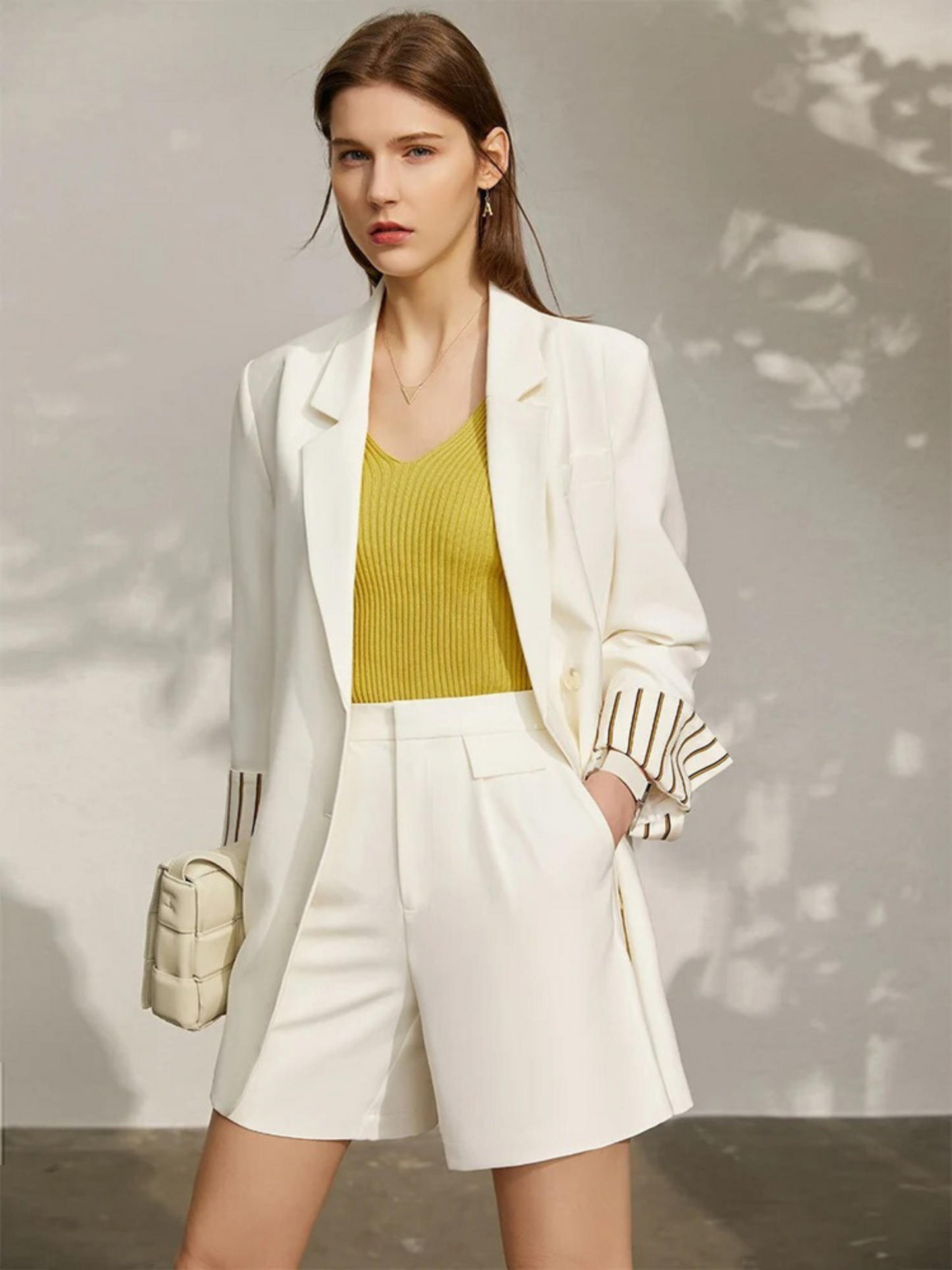 Woman wearing a white blazer, yellow top, and white shorts with a neutral background