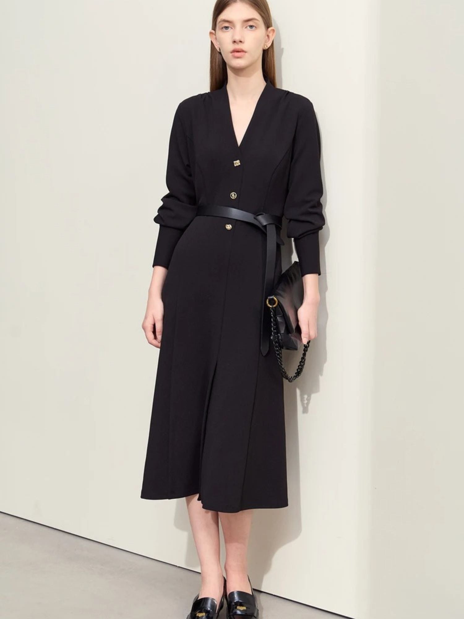 Women's black midi dress with V-neckline, long sleeves, button-front closure, and belted waist. Model wearing black loafers and carrying a black handbag