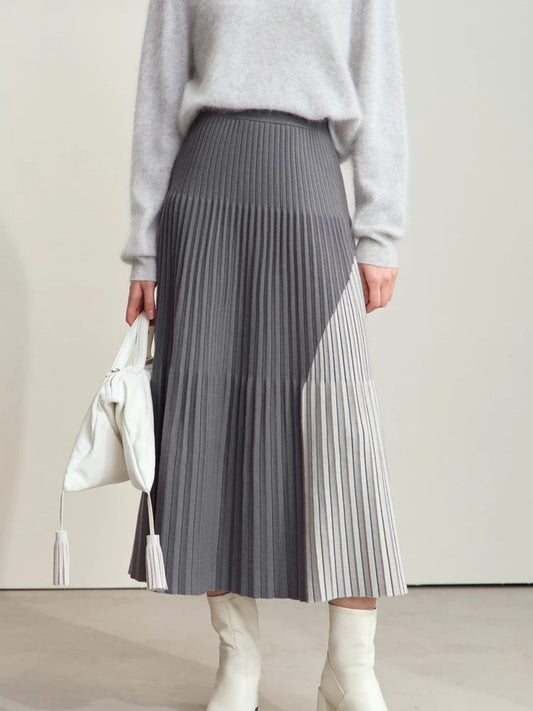 Women's gray pleated midi skirt with color-block design featuring light gray and dark gray panels. Model paired with light gray sweater and white boots