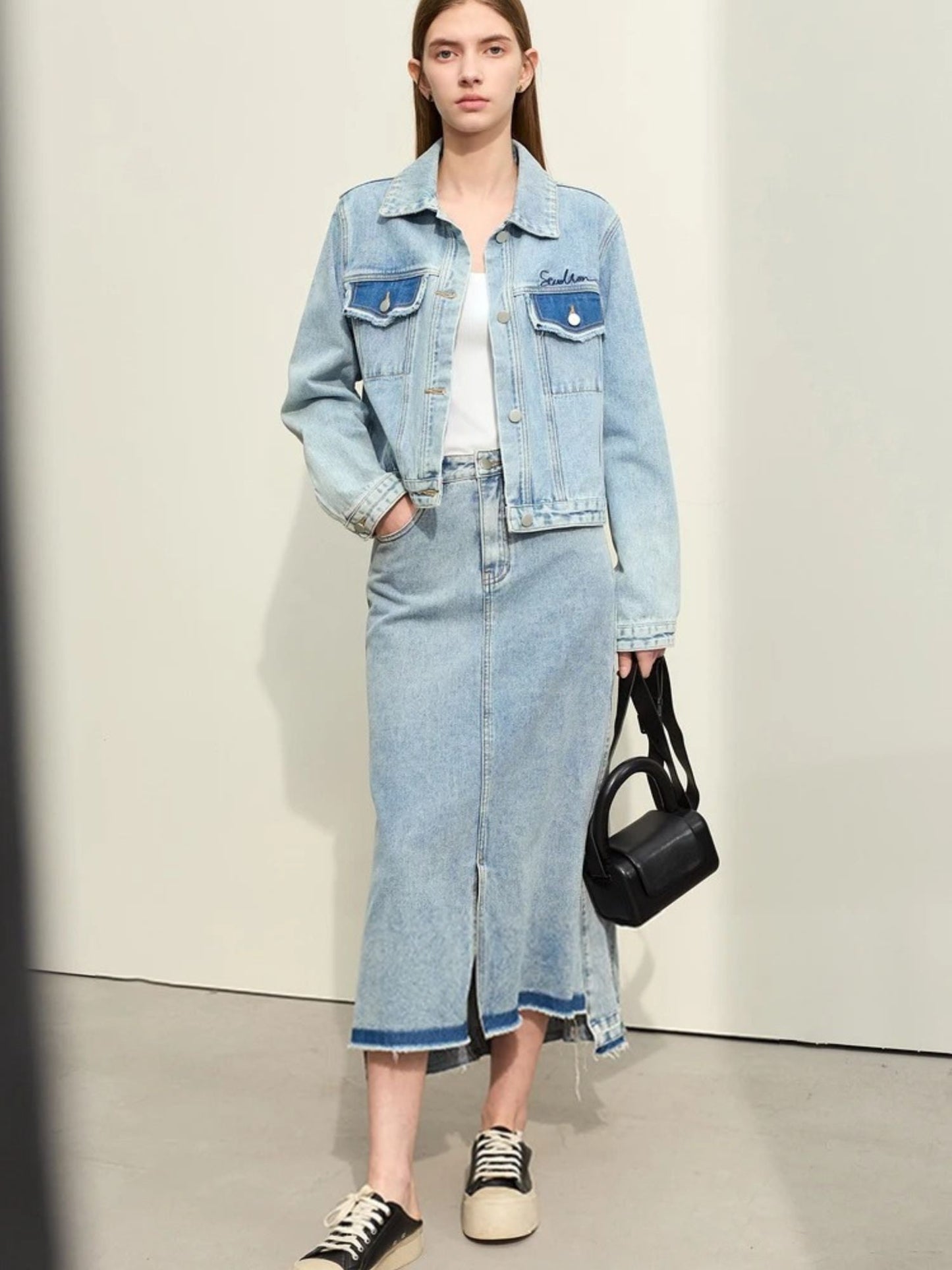 Model in full denim outfit featuring light wash midi skirt with front slit and matching denim jacket. Styled with a black handbag and white flats