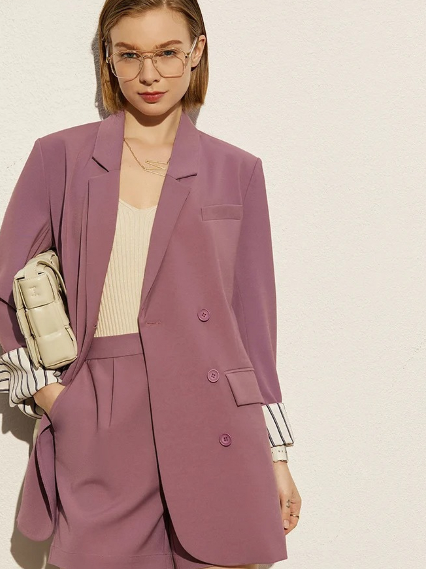 Woman wearing a mauve suit with a beige top and holding a beige bag on a light background