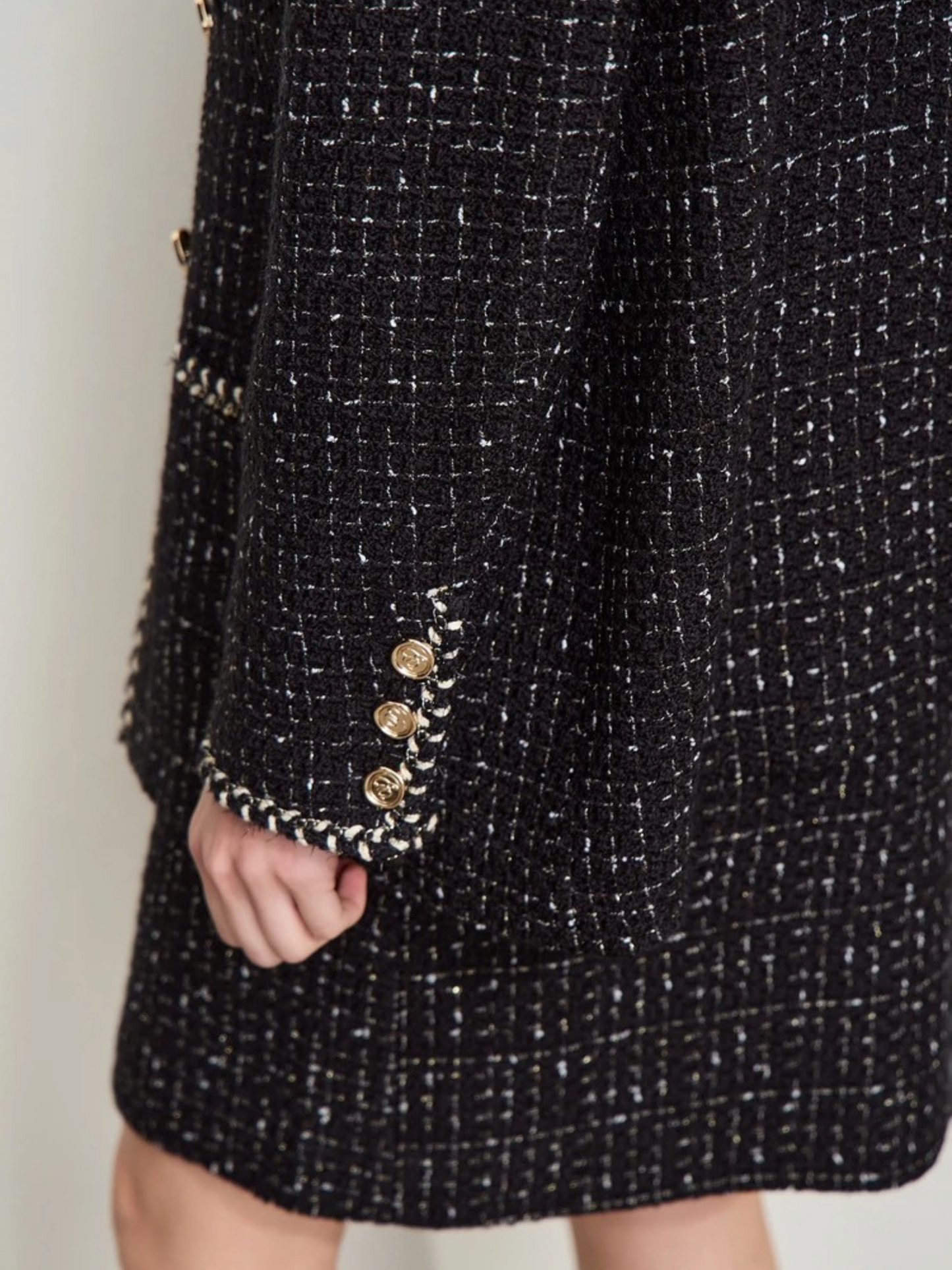 This image shows a close-up of a woman's hand resting near the hem of a black tweed jacket and matching skirt. The fabric features a textured checkered pattern with white and metallic threads woven throughout. The sleeve of the jacket has four decorative gold buttons arranged vertically near the cuff, and the edges are trimmed with a black and white braided detail. The overall look is elegant and sophisticated, highlighting the craftsmanship of the garment