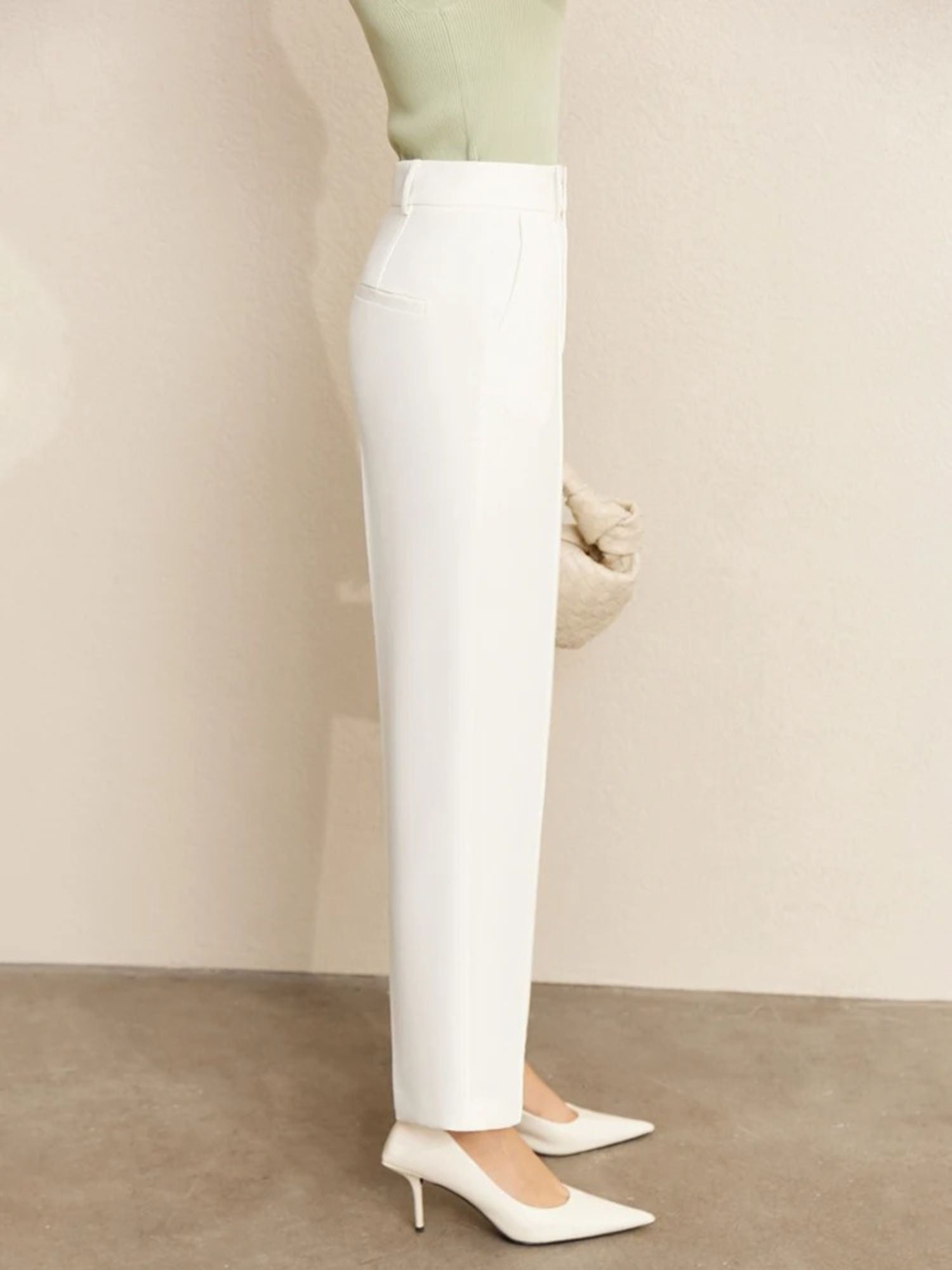 Close-up side view of high-waisted white trousers with tucked-in green top and beige handbag