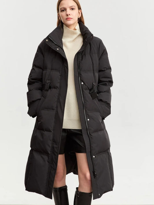 A female model stands front-facing wearing a long black padded winter coat over a beige turtleneck sweater and black shorts. The coat is mid-calf length, with a hood and large front pockets. She is styled with knee-high black boots