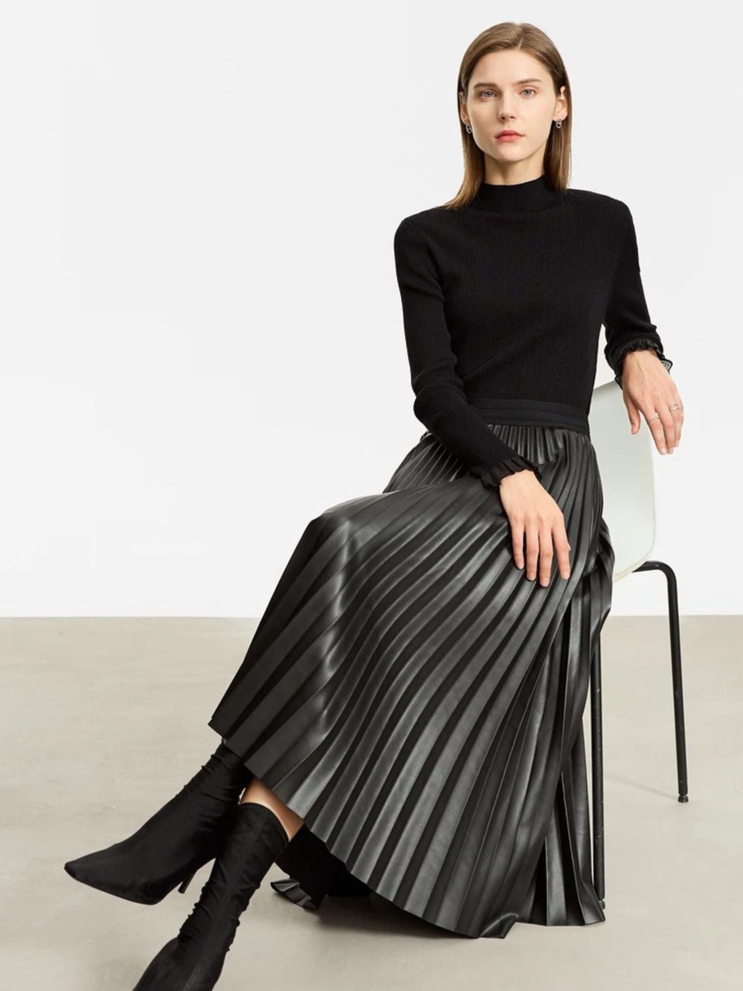 Side view of black faux leather pleated midi skirt worn with black long-sleeve top, model seated