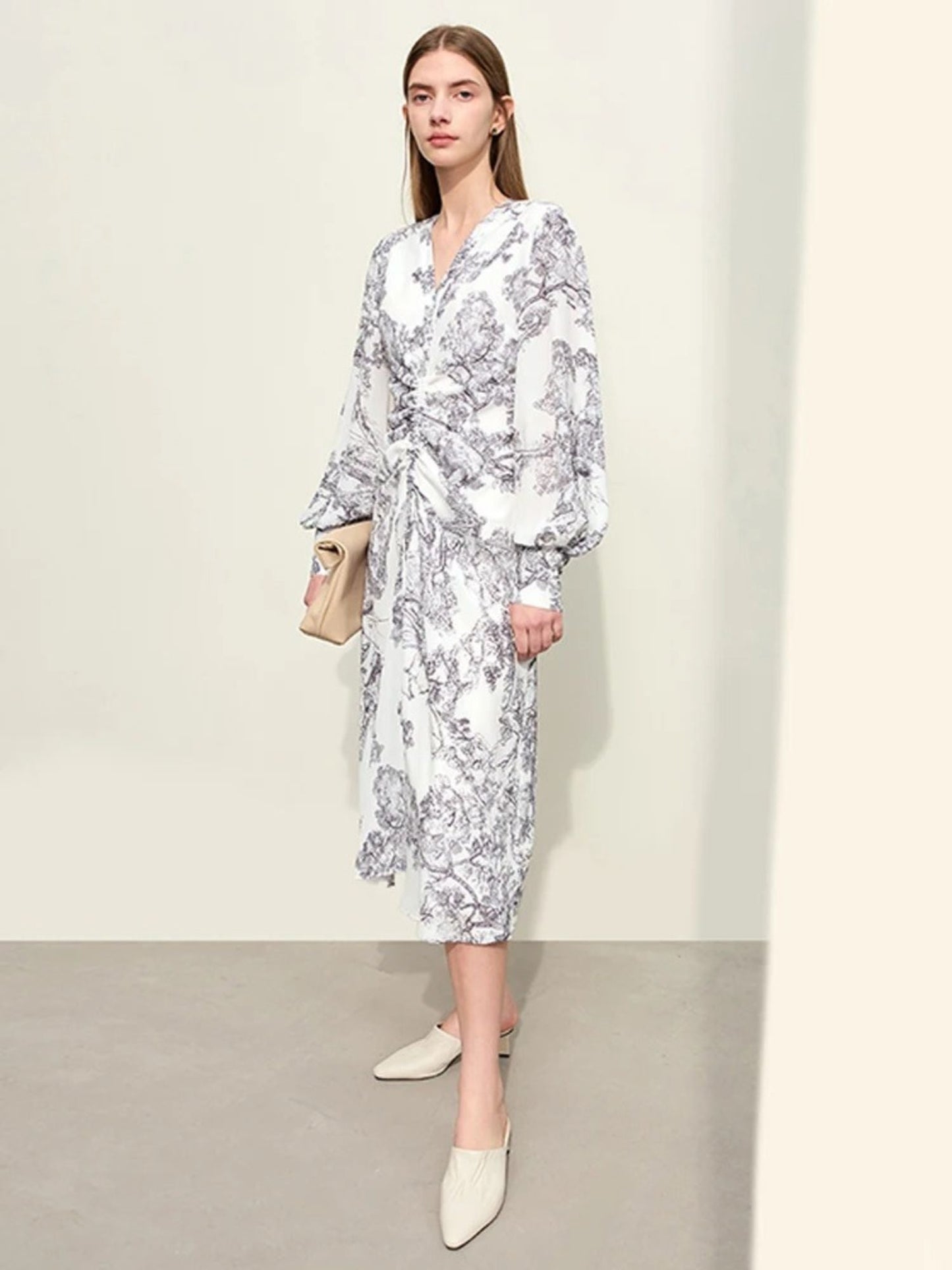 White long-sleeve wrap midi dress with blue floral print, ruched front, and V-neckline. Model walking, holding beige handbag