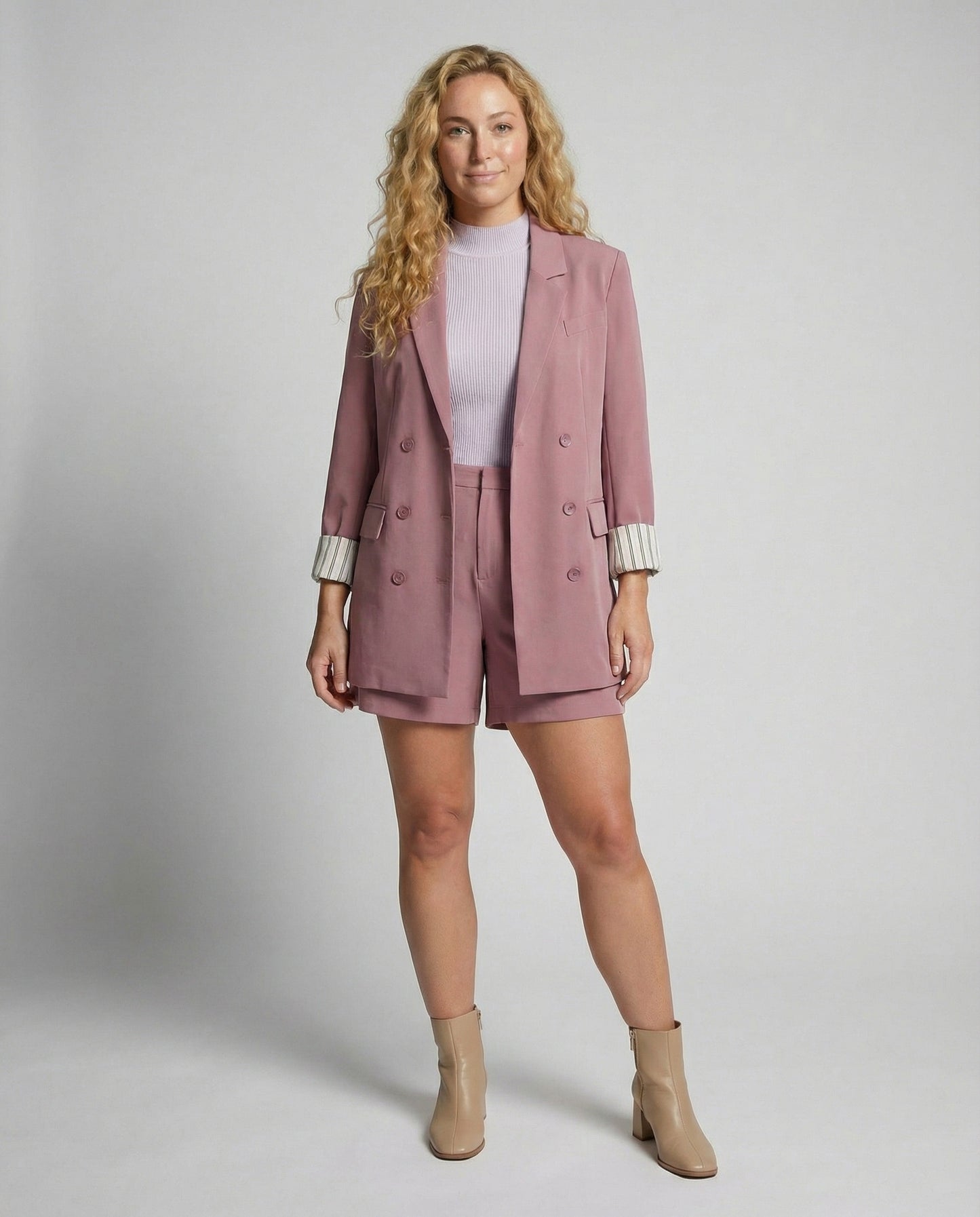 Woman wearing a pink blazer and shorts set with beige boots on a plain background